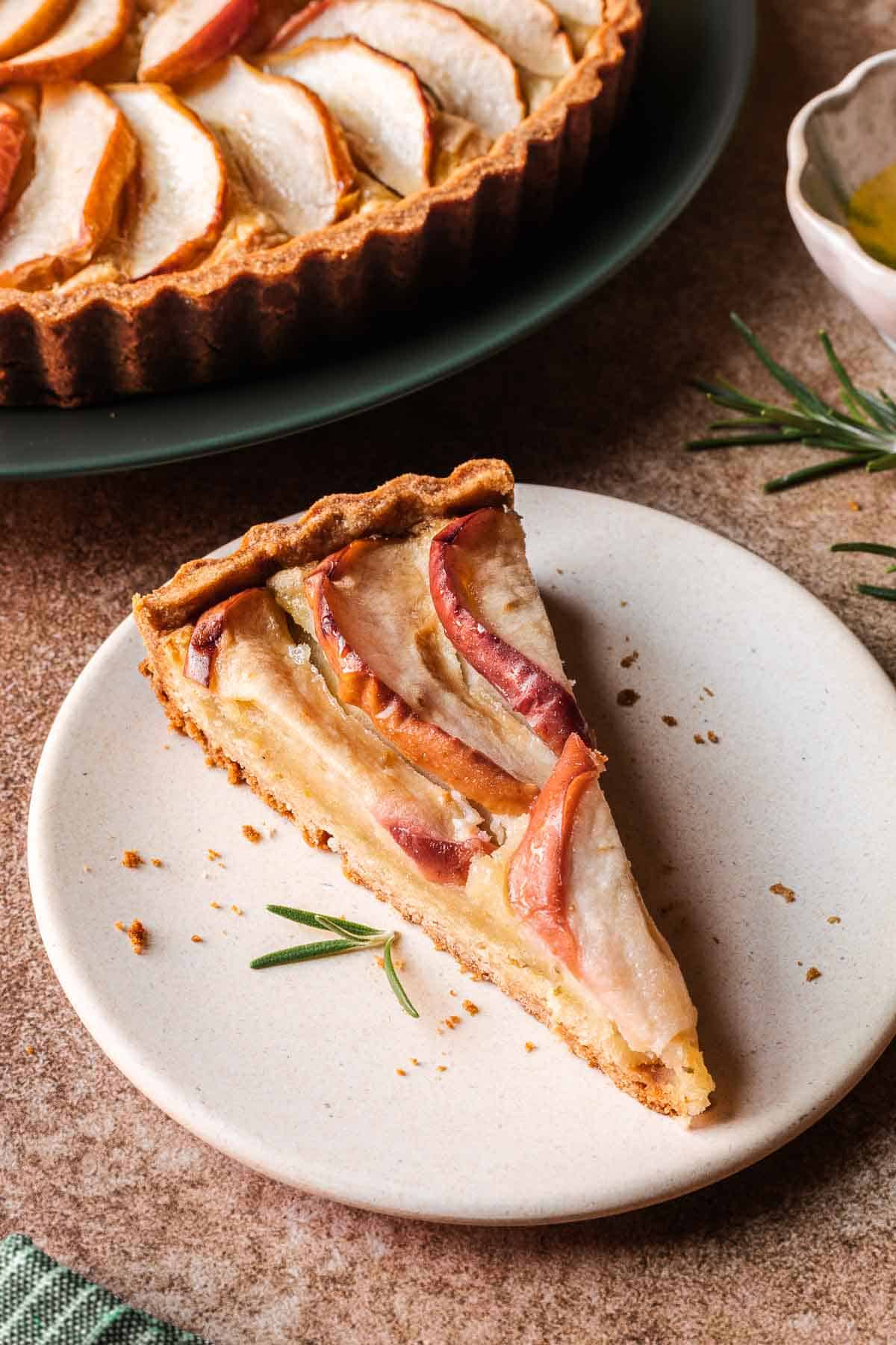 A closeup of a single piece of apple tart on a plate, with the larger tart in the background.
