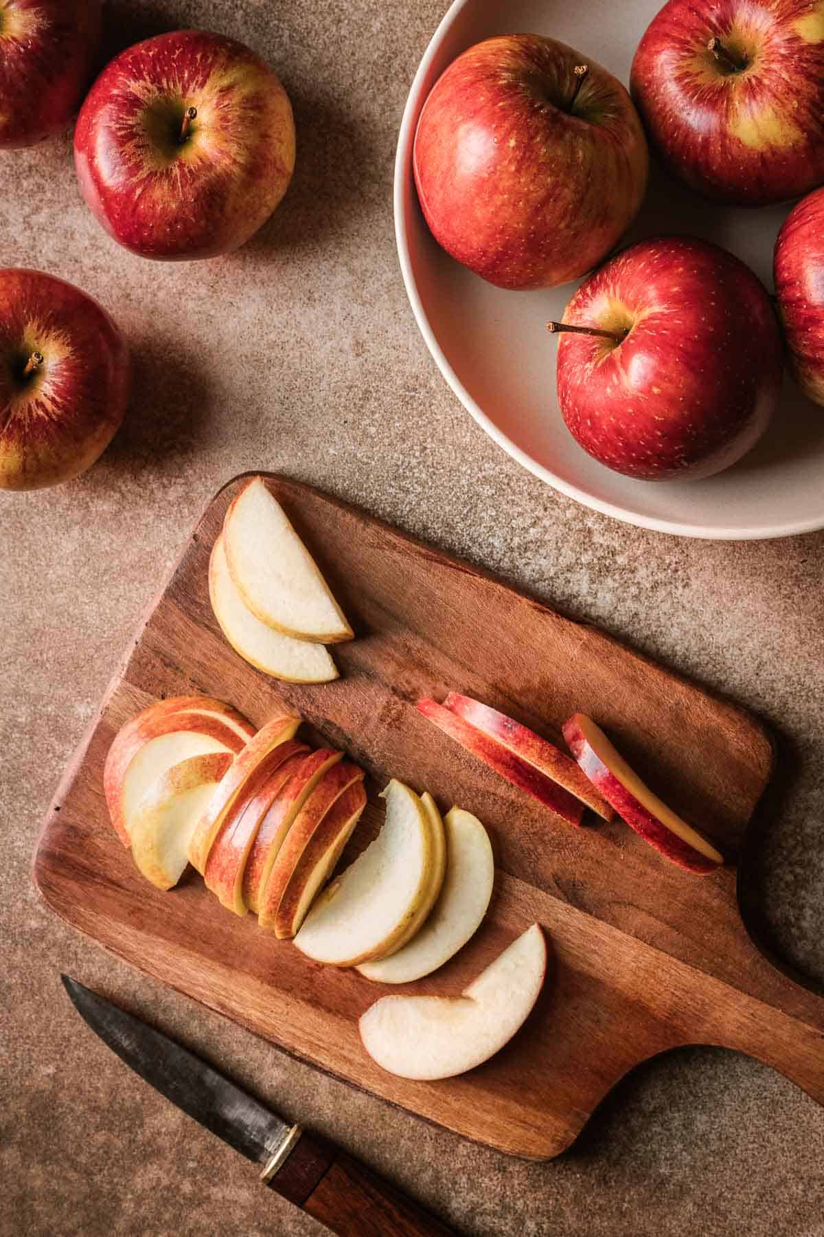 A wooden cutting board with cored, sliced red apples and a bowl of apples nearby.