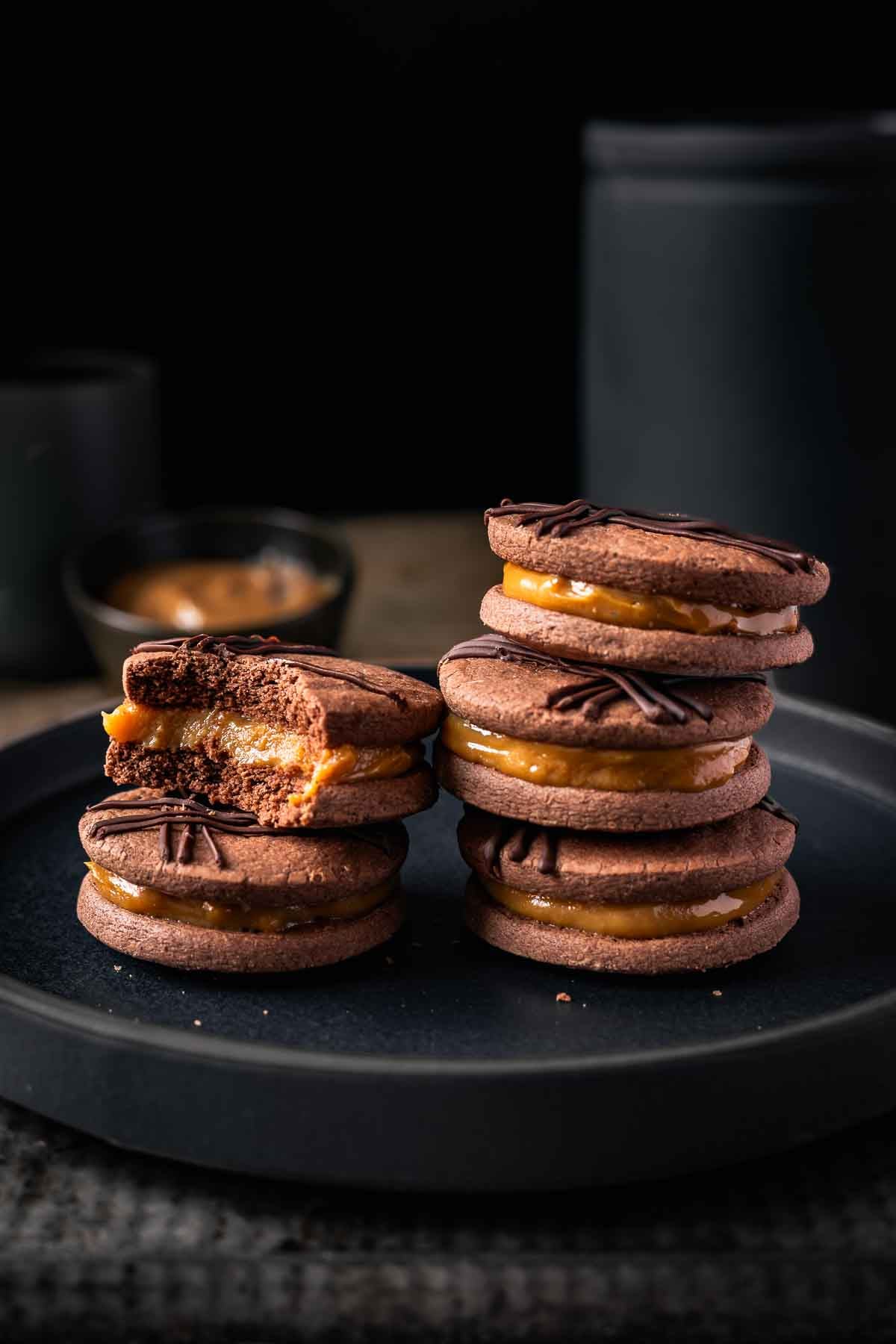 Two stacks of chocolate alfajores with a bite taken out of one cookie showing the dulce de leche center.